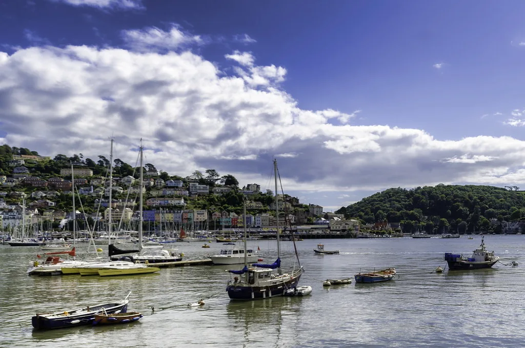 A sunny South Devon estuary with sailing boats moored in front of a hillside of pastel houses